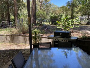 Un patio al aire libre con una mesa y sillas de vidrio, rodeado de un jardín y una parrilla.