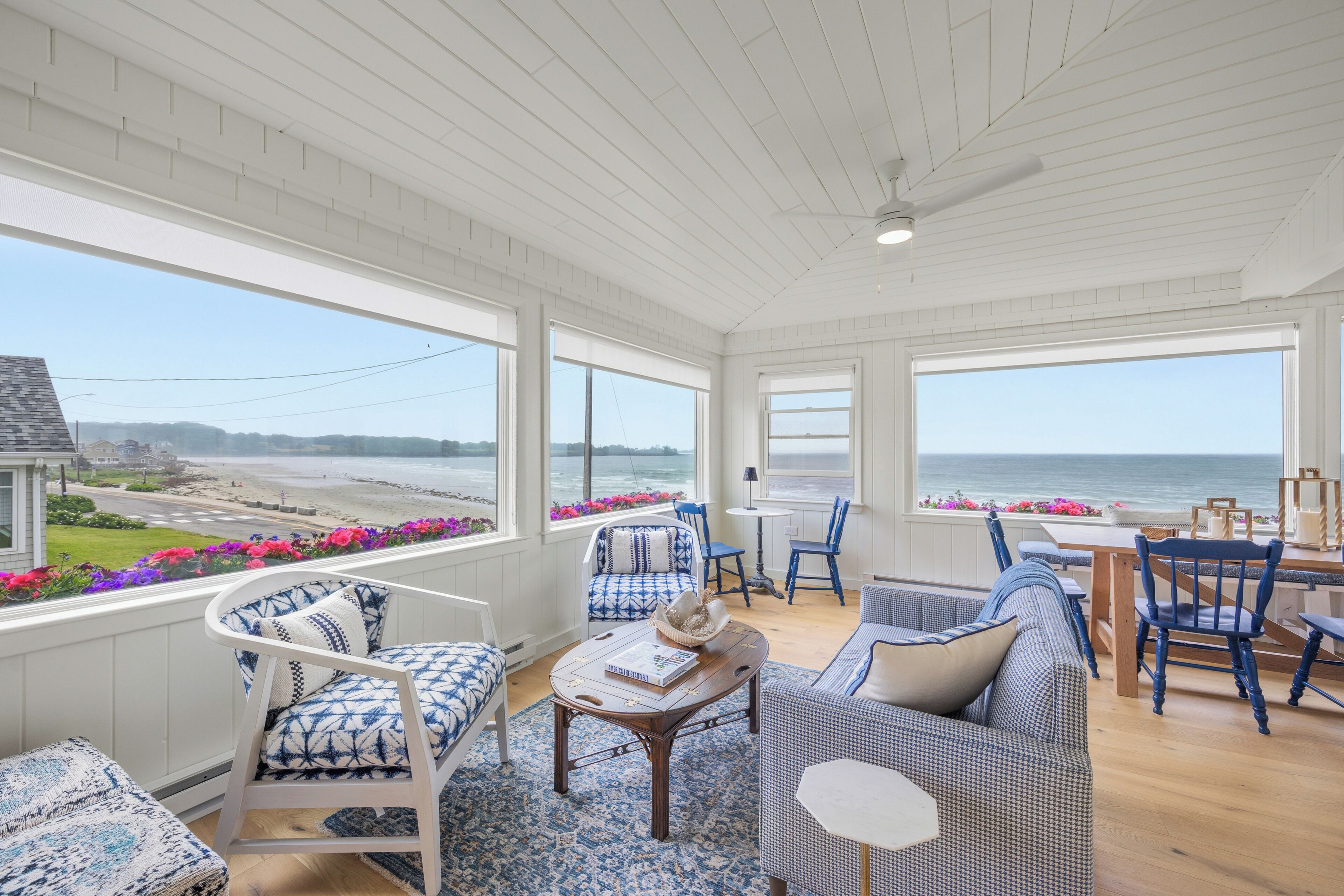 A spacious room with large windows overlooking a beach, furnished with blue and white patterned chairs and a wooden table.