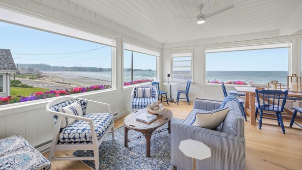 Une pièce spacieuse avec de grandes fenêtres donnant sur une plage, meublée de chaises à motifs bleus et blancs et d’une table en bois.