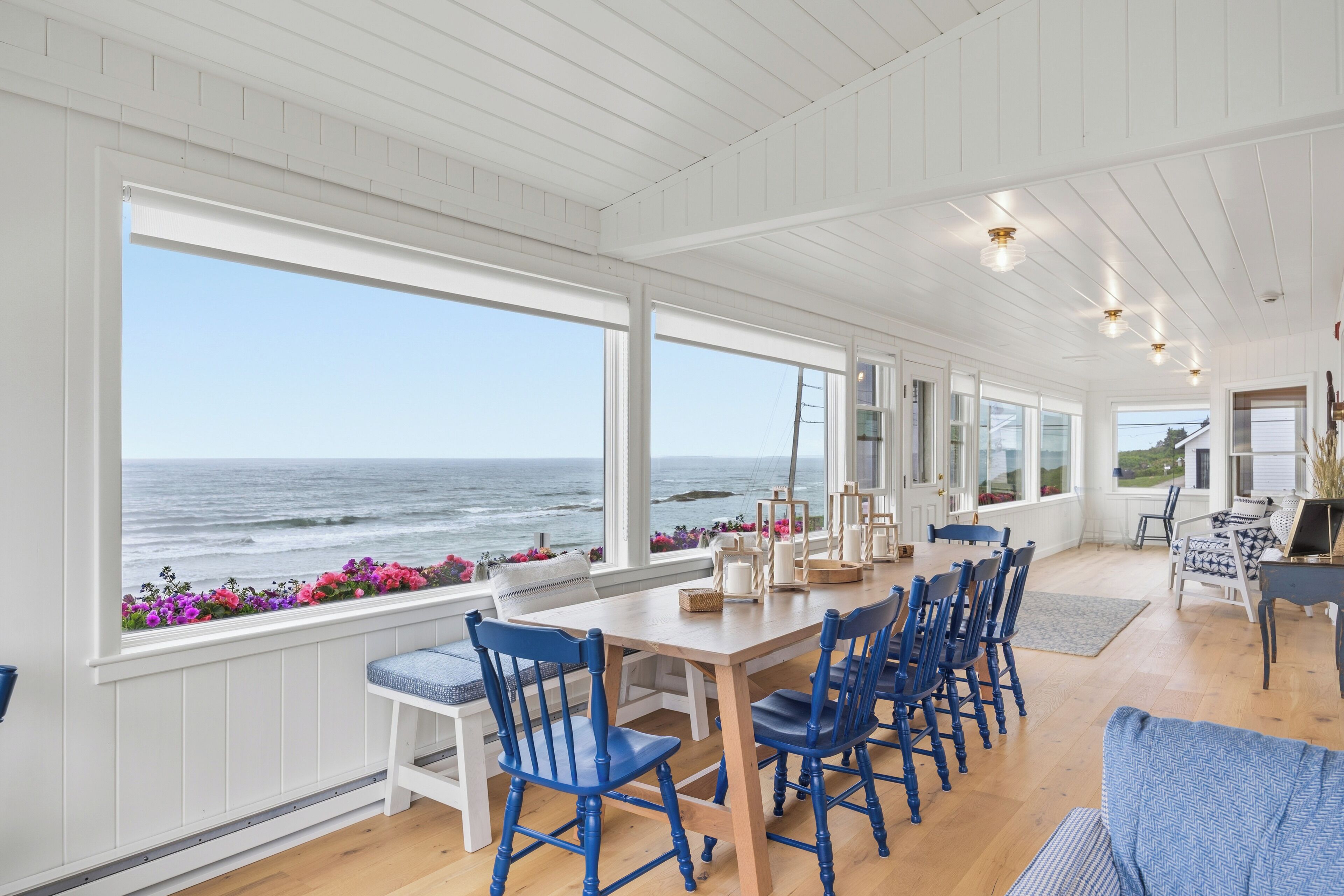 A coastal room with a large window overlooking the ocean, a wooden dining table with blue chairs, and a view of the sea.