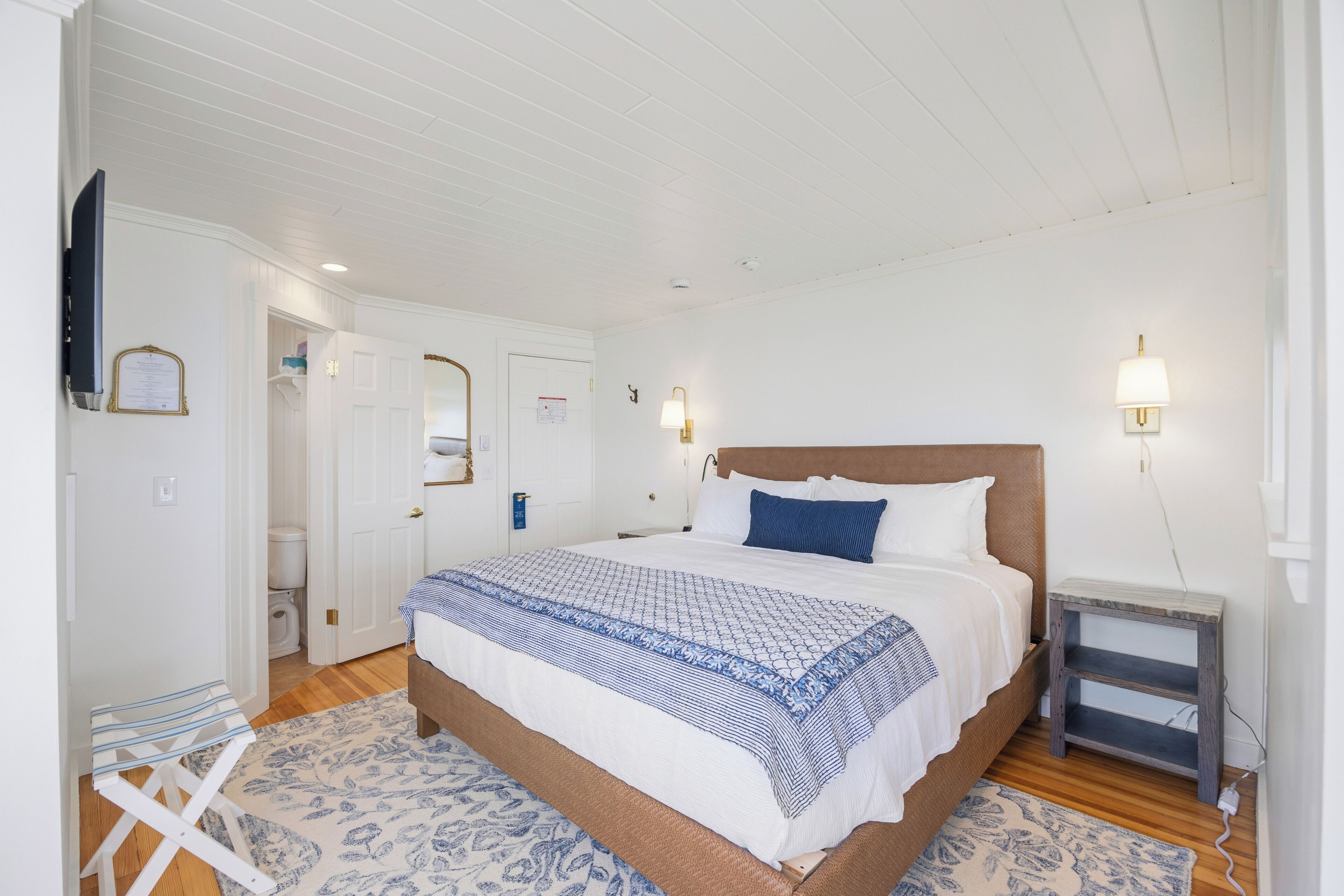 Une chambre à coucher comprenant un lit, des tables de chevet, une commode, un miroir, une porte et un téléviseur fixé au mur.