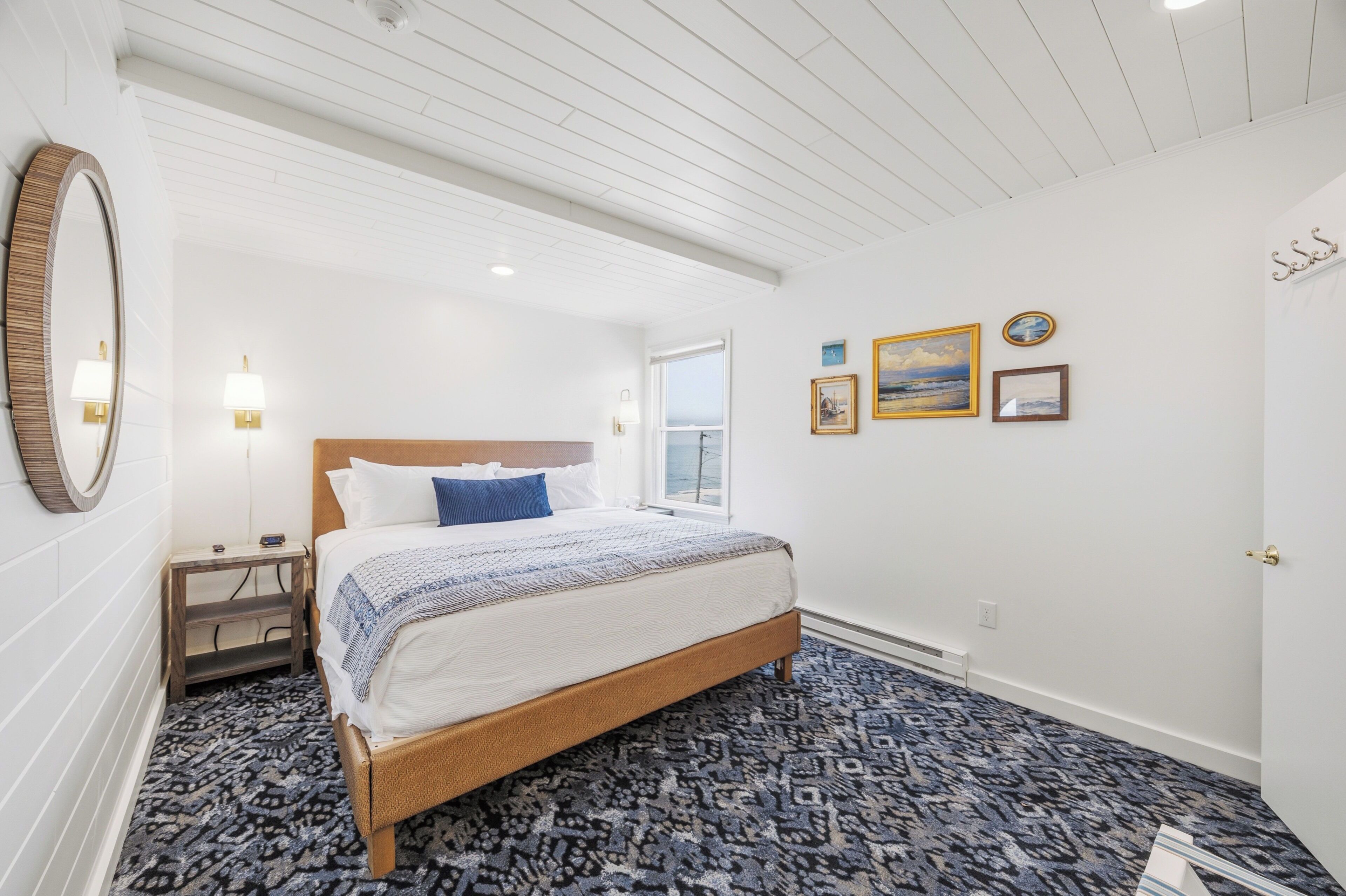 A bedroom with a bed, a nightstand, a wall-mounted mirror, a window, and framed pictures on the wall.