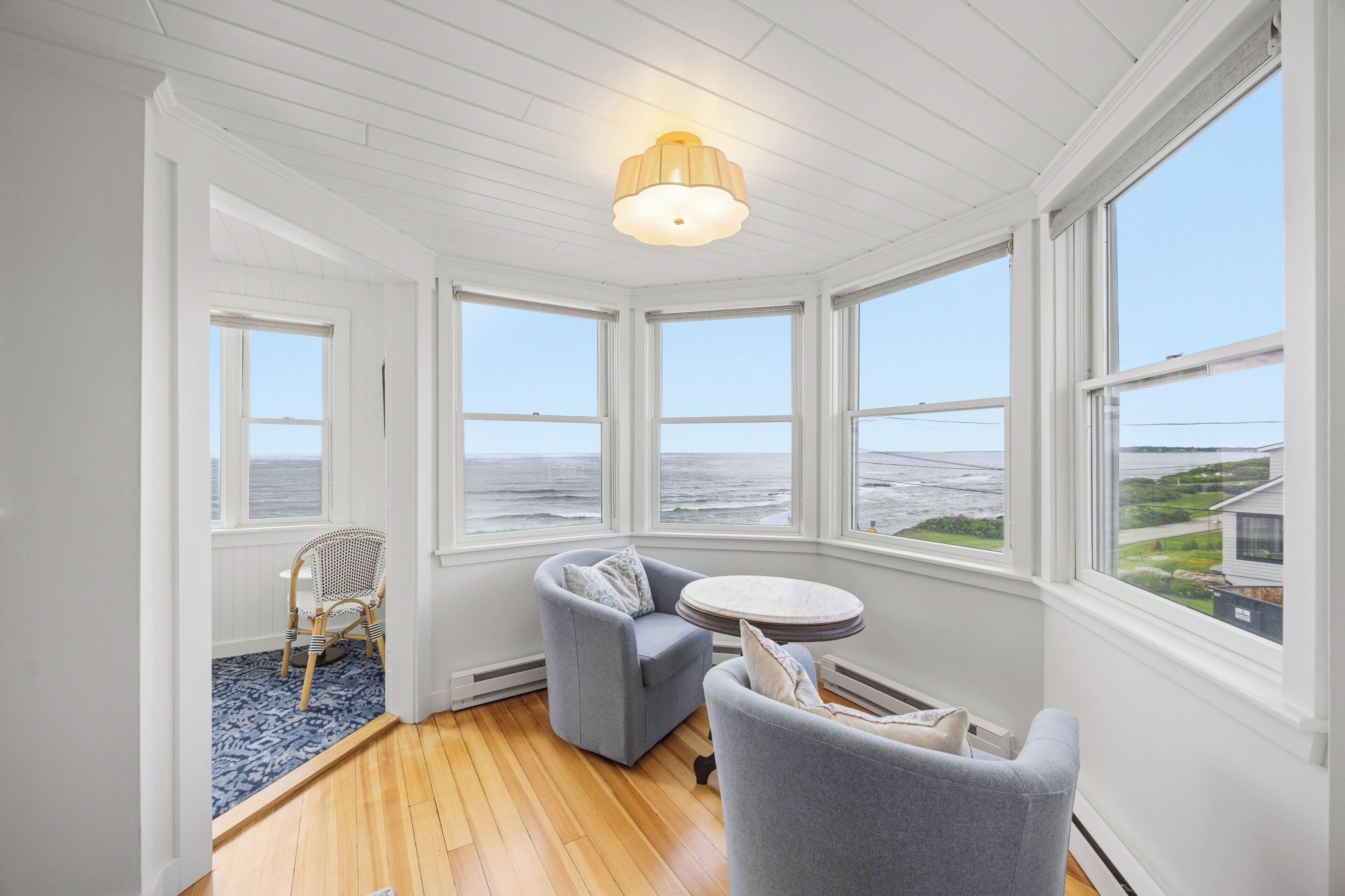 Ein Zimmer mit Holzboden, weißen Wänden und großen Fenstern mit Blick auf das Meer. Zwei graue Sessel und ein kleiner runder Tisch mit einer Lampe stehen dort.