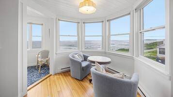 Quarto com piso de madeira, paredes brancas e grandes janelas com vista para o mar. Há duas poltronas cinzas e uma mesa redonda pequena com um abajur.