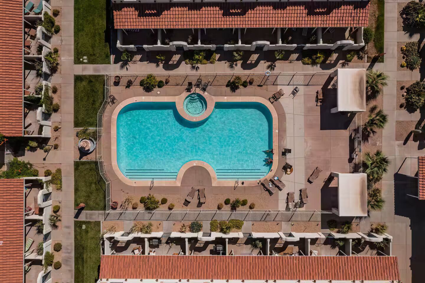An aerial view of a hotel with a central swimming pool, lounge areas, and a covered patio.