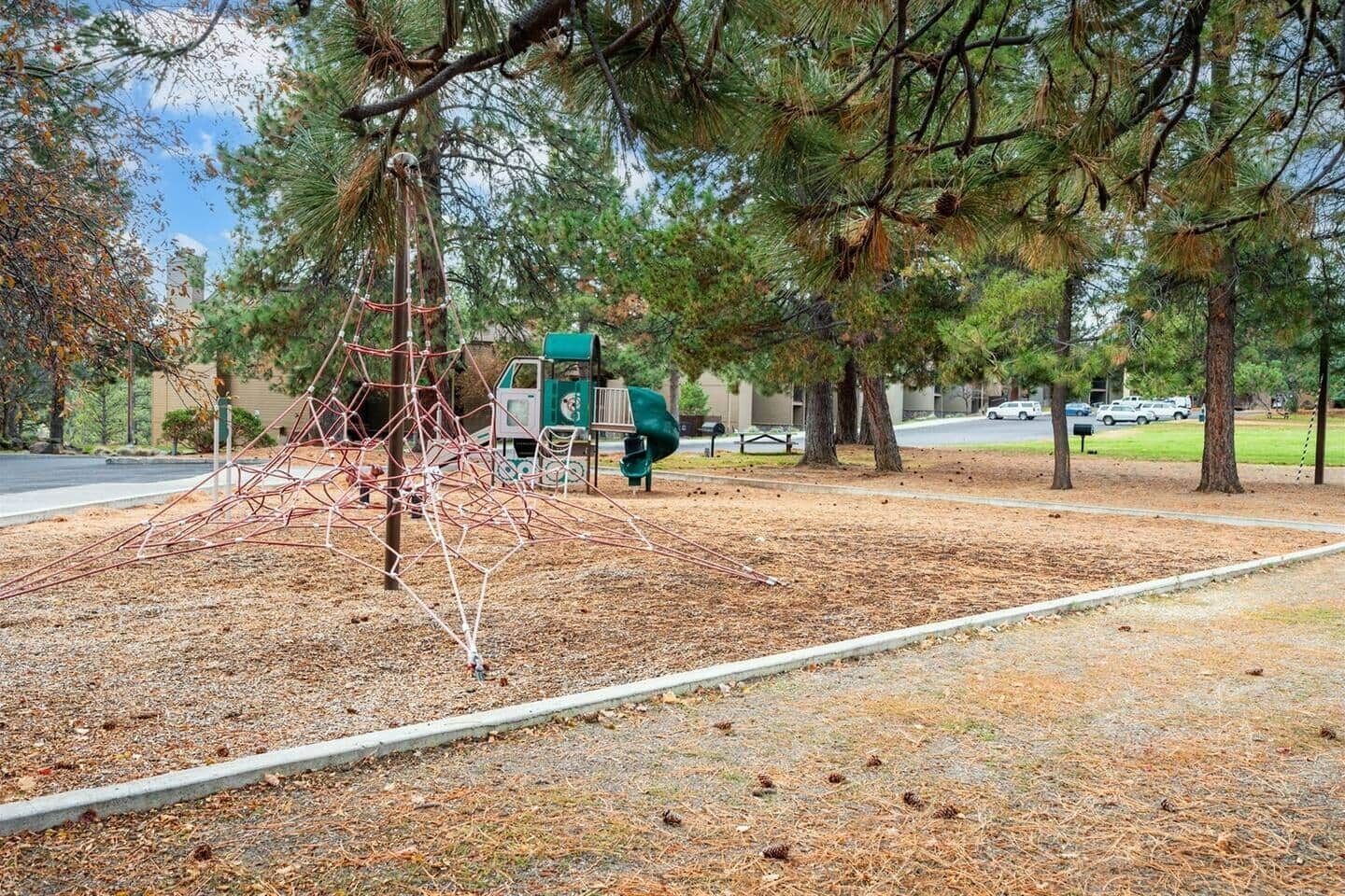Une aire de jeux avec une structure d’escalade, une petite maison de jeu verte et des arbres tout autour.