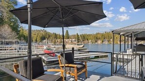 A deck with two chairs and a large umbrella, overlooking a marina with boats and a forested shoreline. - Serene 6-bedroom lake front house in beautiful Lake Arrowhead (Lake Arrowhead)