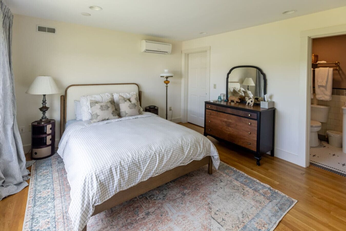 A bedroom with a bed, dresser, mirror, bedside lamp, and a bathroom visible through an open door.
