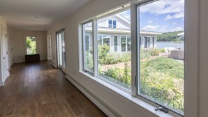 Een lange, smalle kamer met grote ramen, houten vloeren en uitzicht op een huis en tuin.