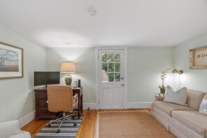 Ein Zimmer mit Schreibtisch, Stuhl, Fernseher, Lampe, Sofa und einer Tür mit Fenster.