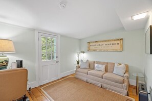 Een kamer met een witte deur, een beige bank, een bord met een lijst aan de muur, en een lamp op een tafel.