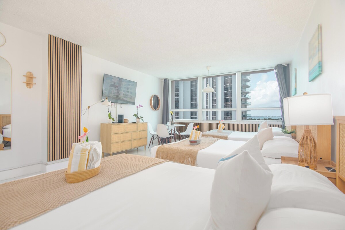 A hotel room with two beds, a TV, a large window with a view, a dresser, and a dining area.