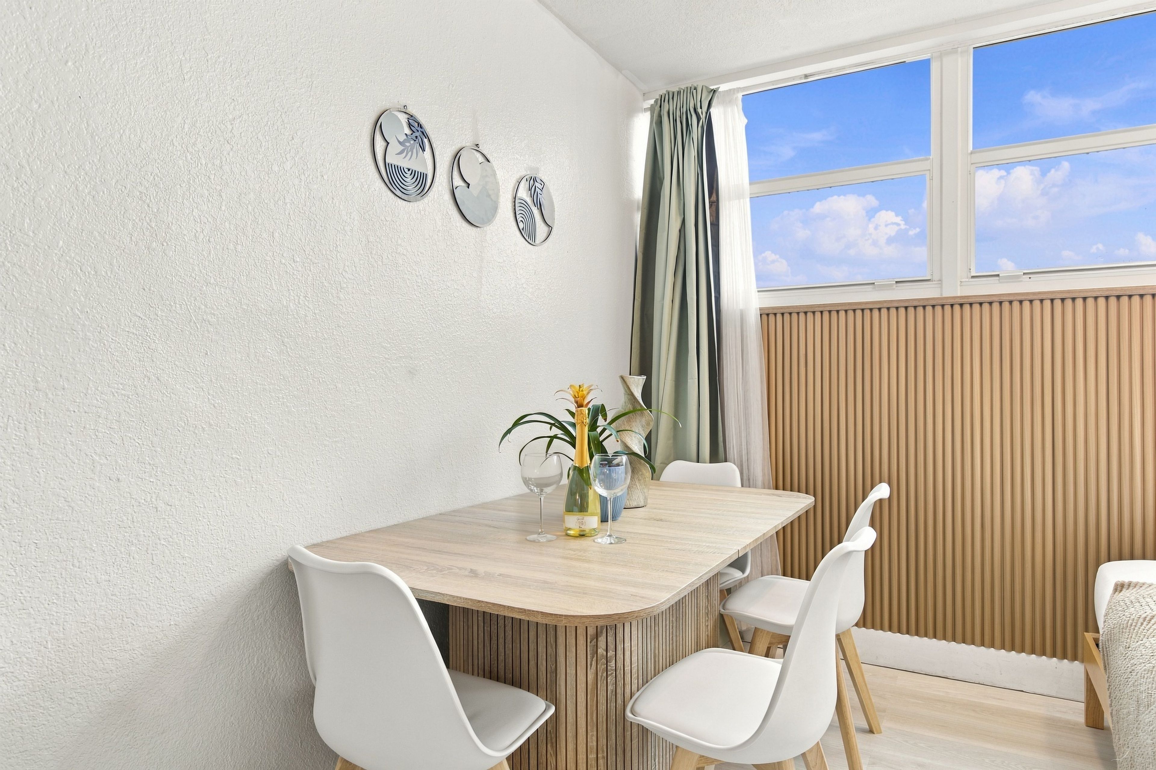 Uma sala de jantar com mesa de madeira, cadeiras brancas, um vaso com flores e uma janela com cortinas.