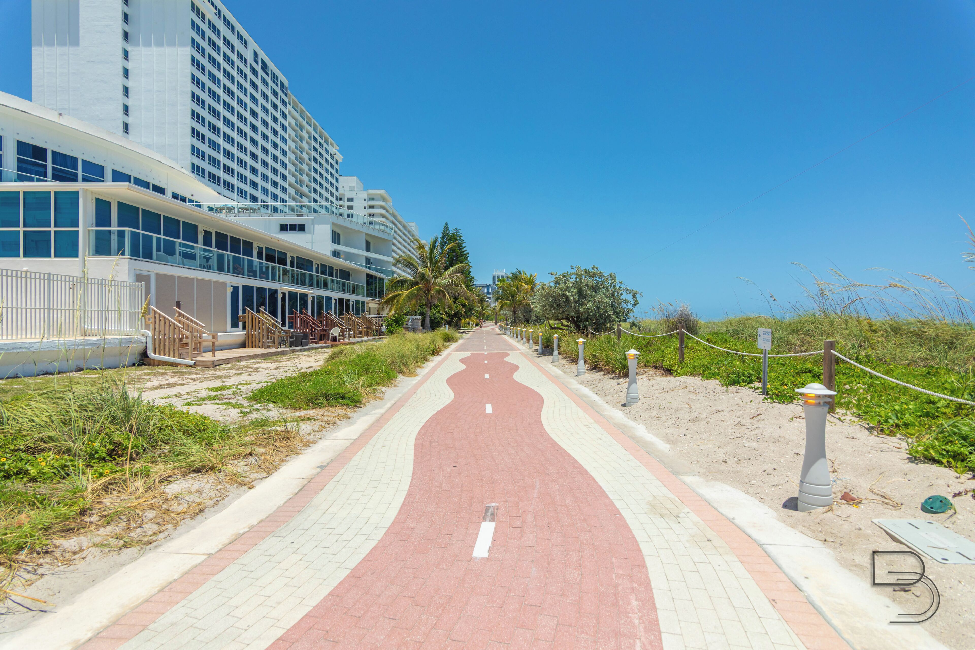 A pathway leading to a hotel with a modern design, featuring a red and white color scheme, and a clear view of the beach and palm trees.