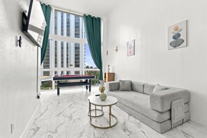 A modern living room with a sofa, a pool table, and a view of a building through large windows. - Boutique Apartments Castle Beach (Miami Beach)