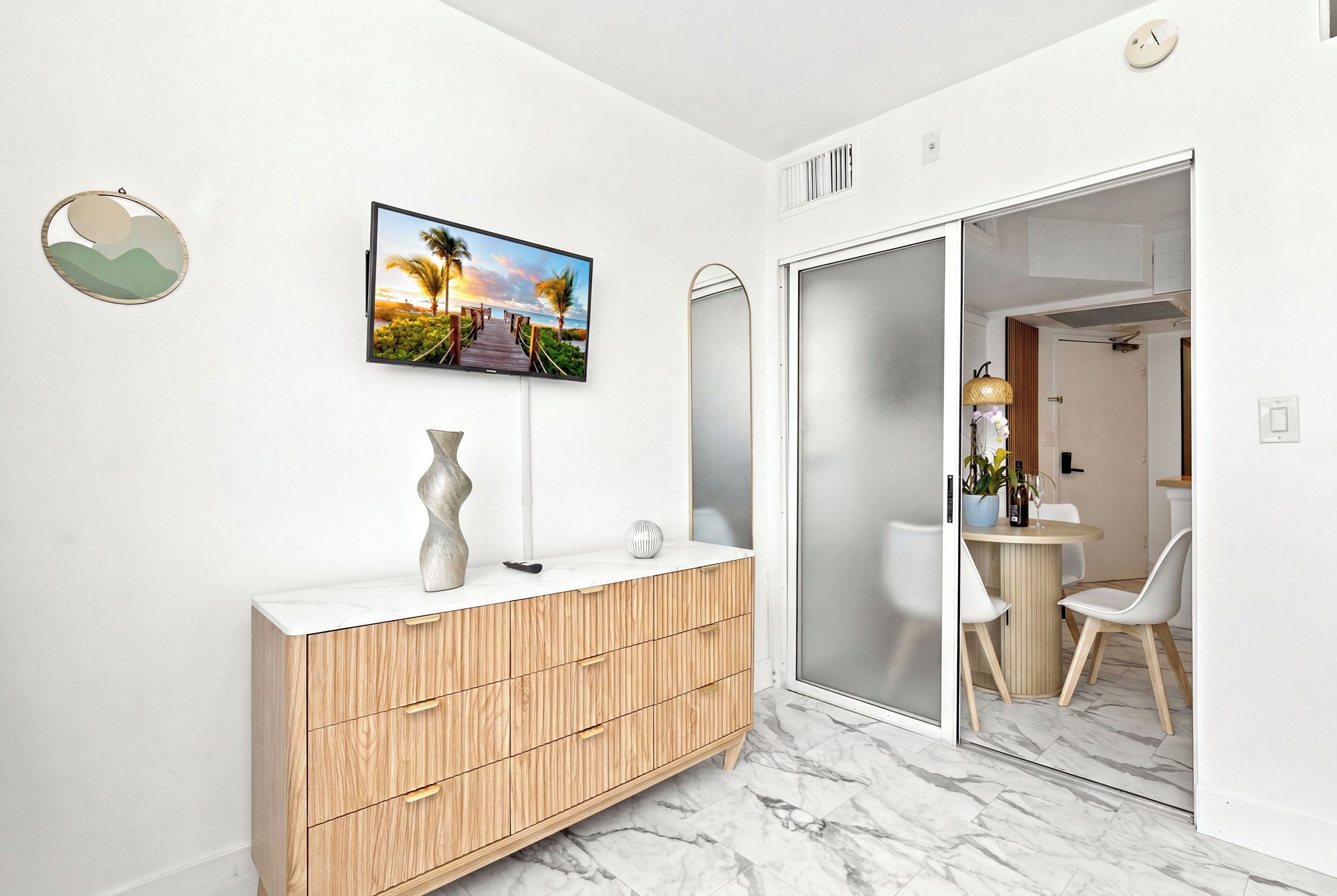 A modern bathroom with a wooden vanity, a large mirror, a wall-mounted TV, and a sliding glass door leading to a dining area.