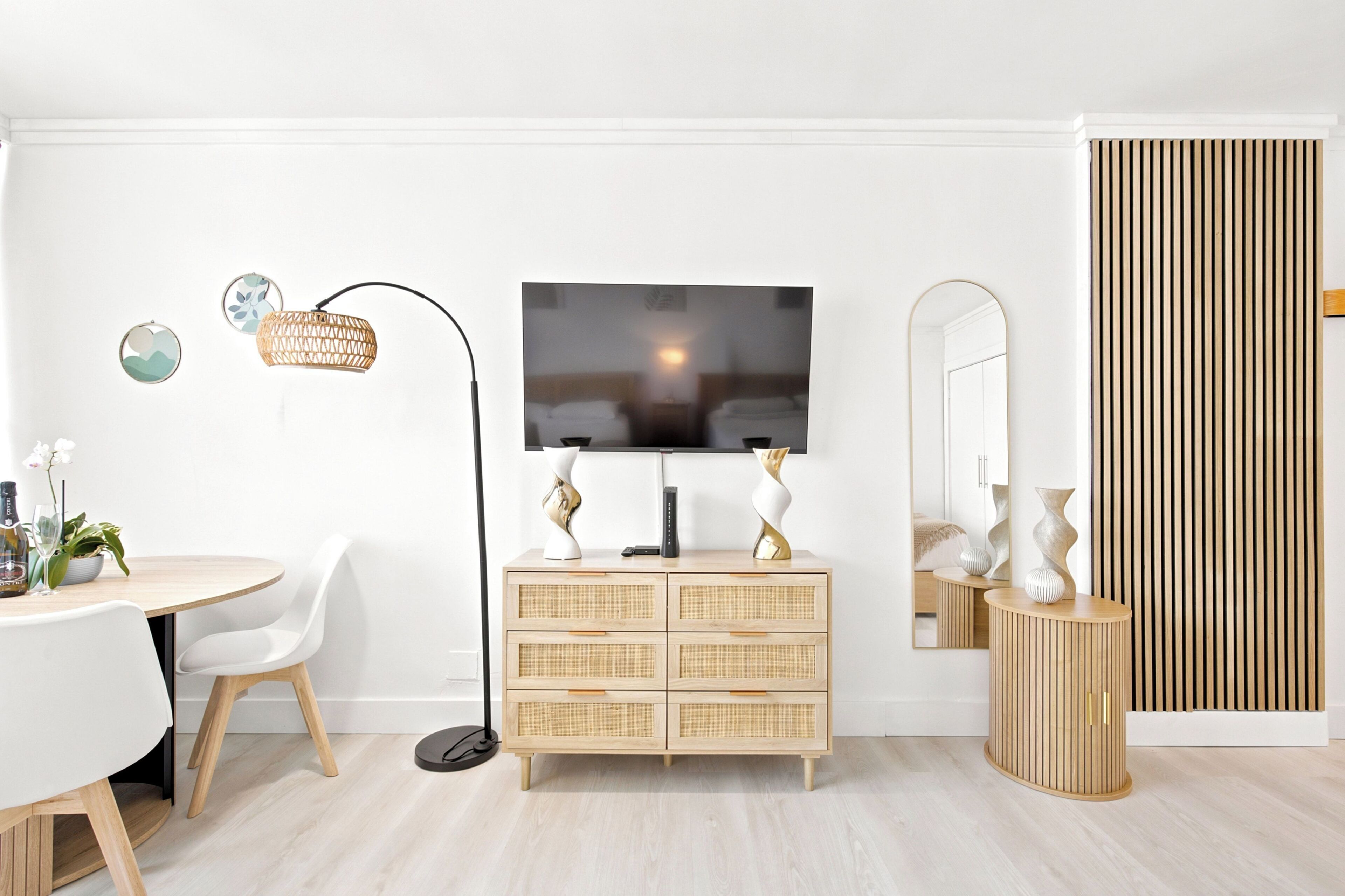 Een moderne woonkamer met een flatscreen-tv, een eettafel met stoelen, een rieten bijzettafeltje, een spiegel en een houten kast.