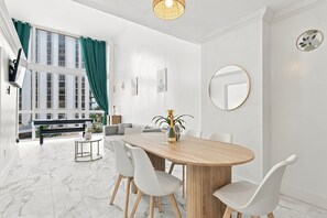 A modern dining area with a wooden table, white chairs, a round mirror, and a view of buildings through large windows. - Boutique Apartments Castle Beach (Miami Beach)