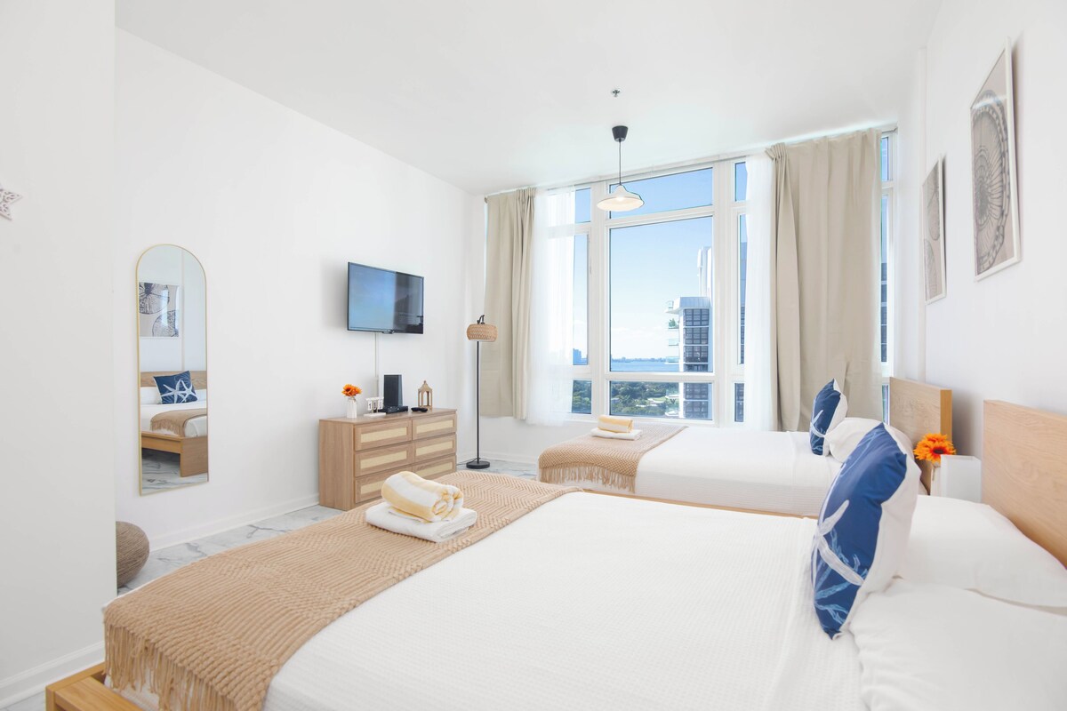 A hotel room with two beds, a TV, a dresser, and a large window with a view.