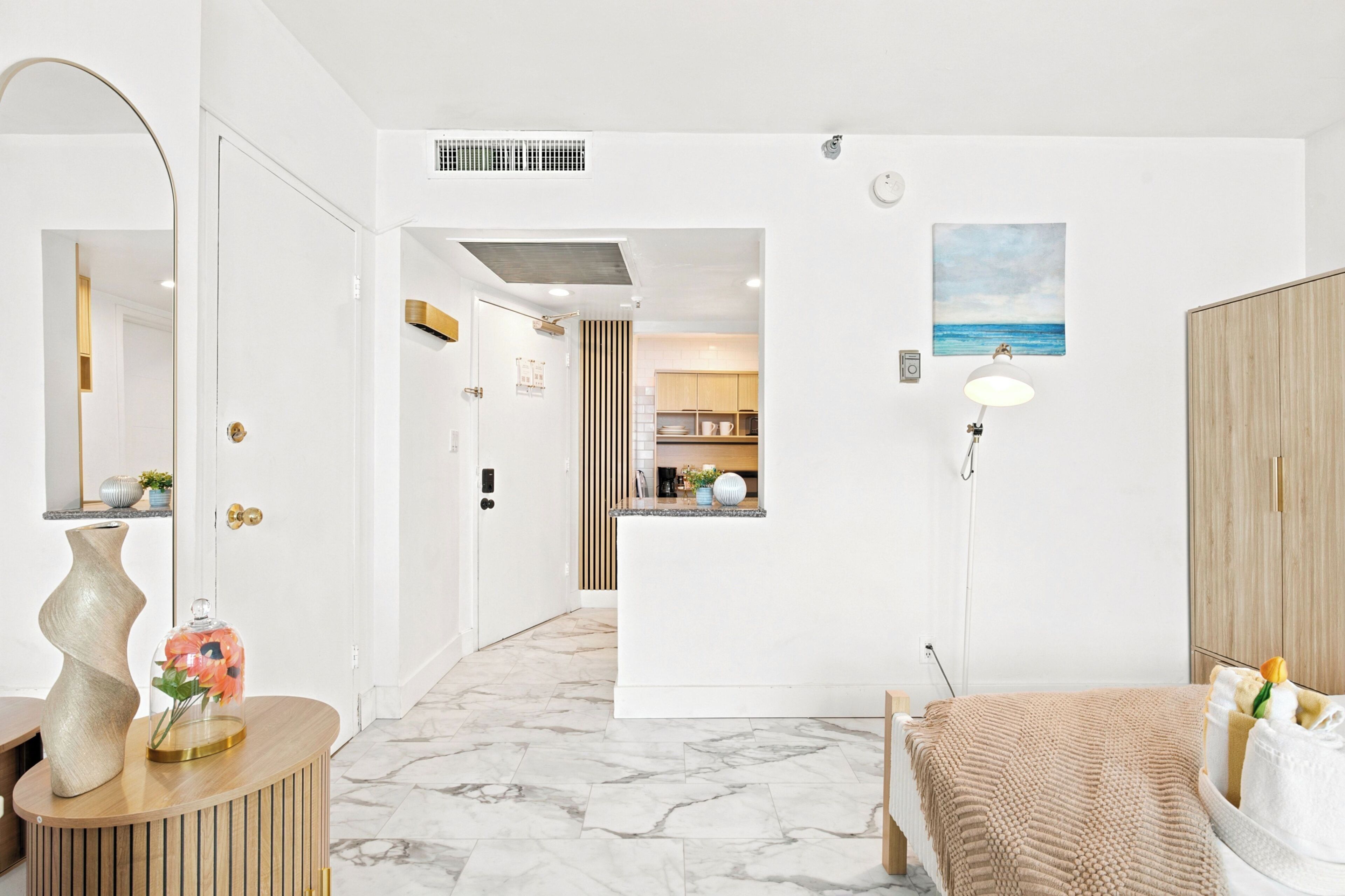 Een hotelkamer met een marmeren vloer, een bed met een beige deken, een bedlamp, een houten kledingkast en uitzicht op de keuken.