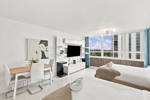 A modern living room with a dining area, a flat-screen TV, and a large window with a city view. - Boutique Apartments Castle Beach (Miami Beach)