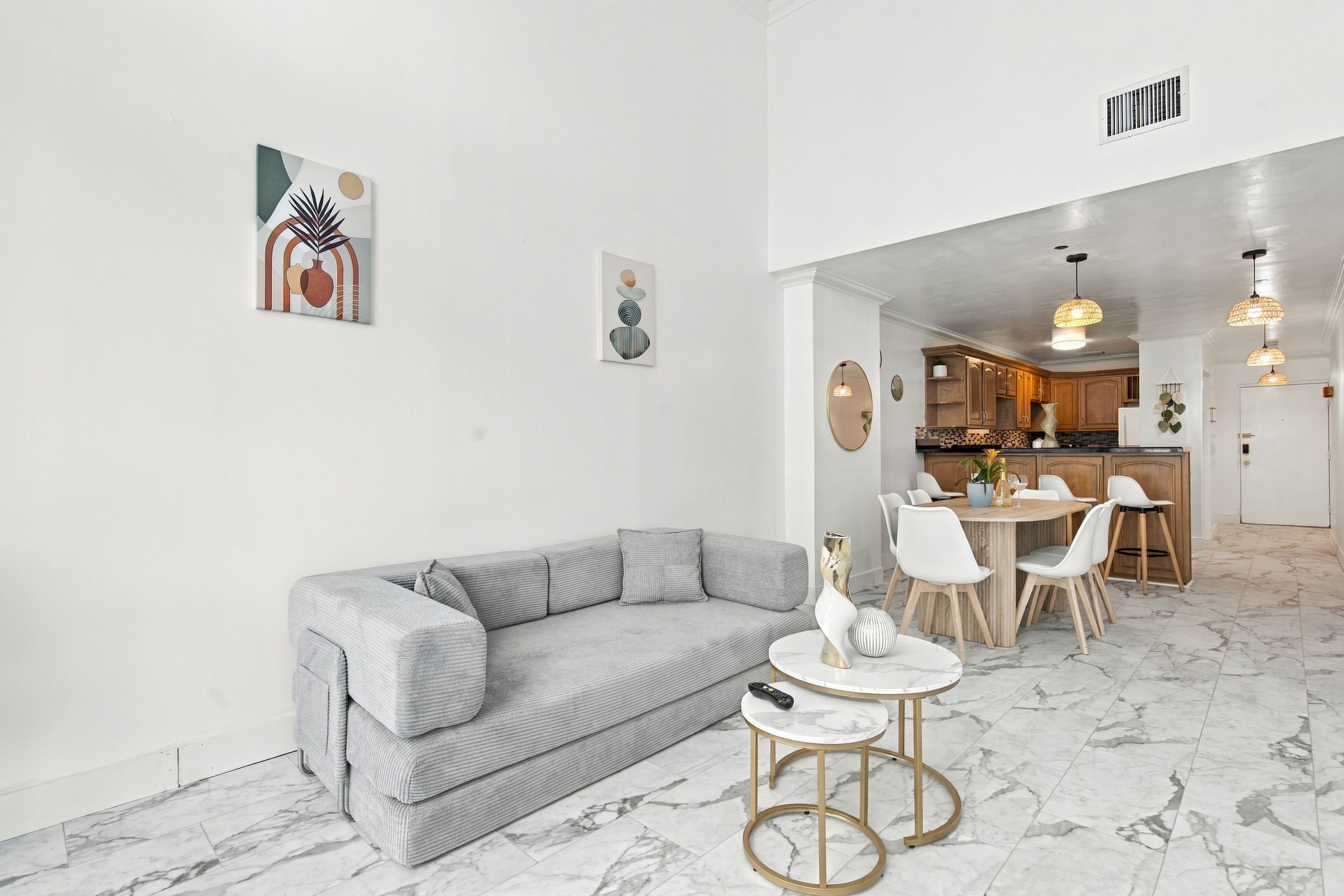 Un salon moderne avec un canapé gris, une petite table ronde ornée d’un vase, et une cuisine en arrière-plan, avec des meubles en bois et un coin repas.