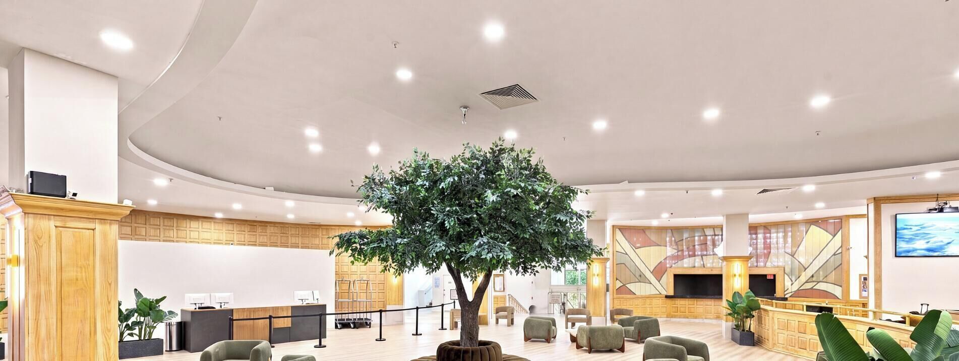 Un vestíbulo de hotel con una recepción, área de descanso, un árbol grande y una pantalla de televisión.
