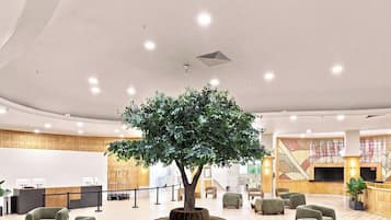 Un vestíbulo de hotel con una recepción, área de descanso, un árbol grande y una pantalla de televisión.