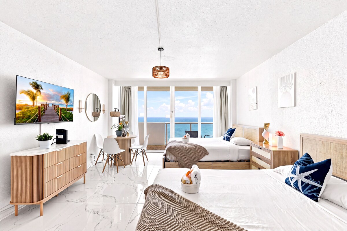 A modern hotel room with a large window, a flat-screen TV, a wooden dresser, a bed with blue and white pillows, and a small table with a vase.