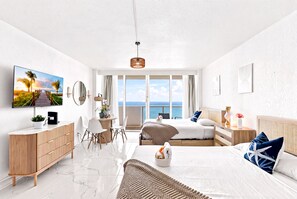 A modern hotel room with a large window, a flat-screen TV, a wooden dresser, a bed with blue and white pillows, and a small table with a vase. - Boutique Apartments Castle Beach (Miami Beach)
