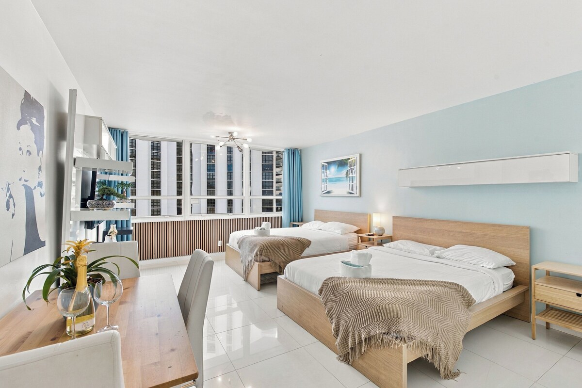 A modern hotel room with two beds, a dining area, and a view of the cityscape.