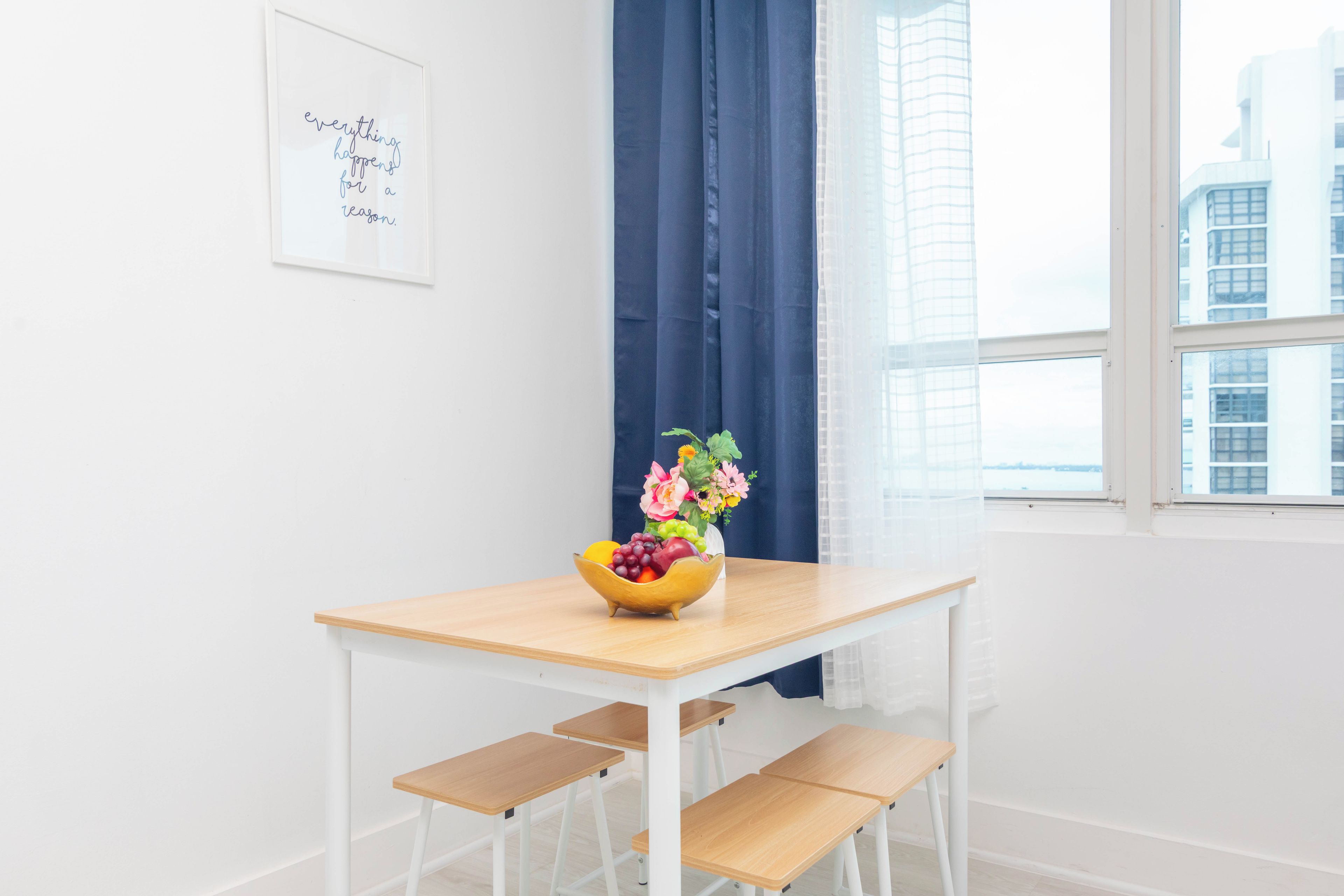 Una habitación minimalista con una mesa de madera, un cuenco de frutas y flores, una cita inspiradora enmarcada y vistas de edificios a través de una ventana.