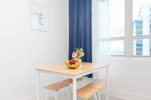 Una habitación minimalista con una mesa de madera, un cuenco de frutas y flores, una cita inspiradora enmarcada y vistas de edificios a través de una ventana.