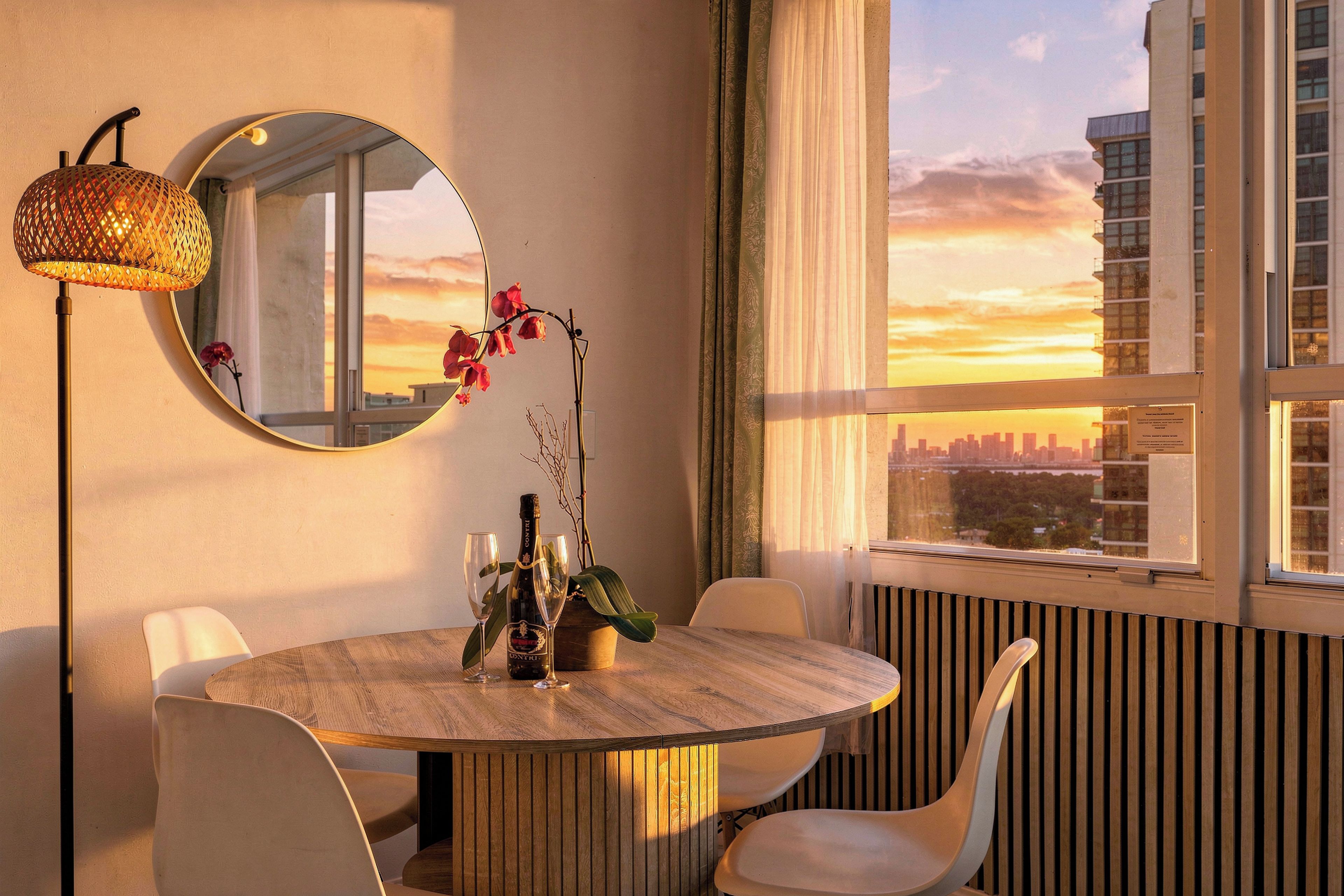 A dining area with a round table, chairs, a mirror, and a view of the city skyline at sunset.