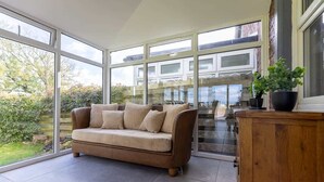 A spacious room with a large window, a beige sofa, and a wooden cabinet. - Spacious 5-Bed Home with Views, Garden & Playroom (Silloth)
