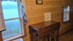 A cabin interior with a wooden wall, a window with a view of a lake, a wooden table, a trash can, and a sign reading "Cozy Cabin #1". - Popeye and Grammy’s Lookout Point: Cozy Lakeside Cabin for Two (Cornell)