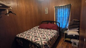 A bedroom with a bed, wooden walls, a ceiling fan, a window with blue curtains, and a small shelf with pillows. - Bob’s and Ella’s Cozy Cabin — Charming Retreat for 6 with Fire Ring (Cornell)