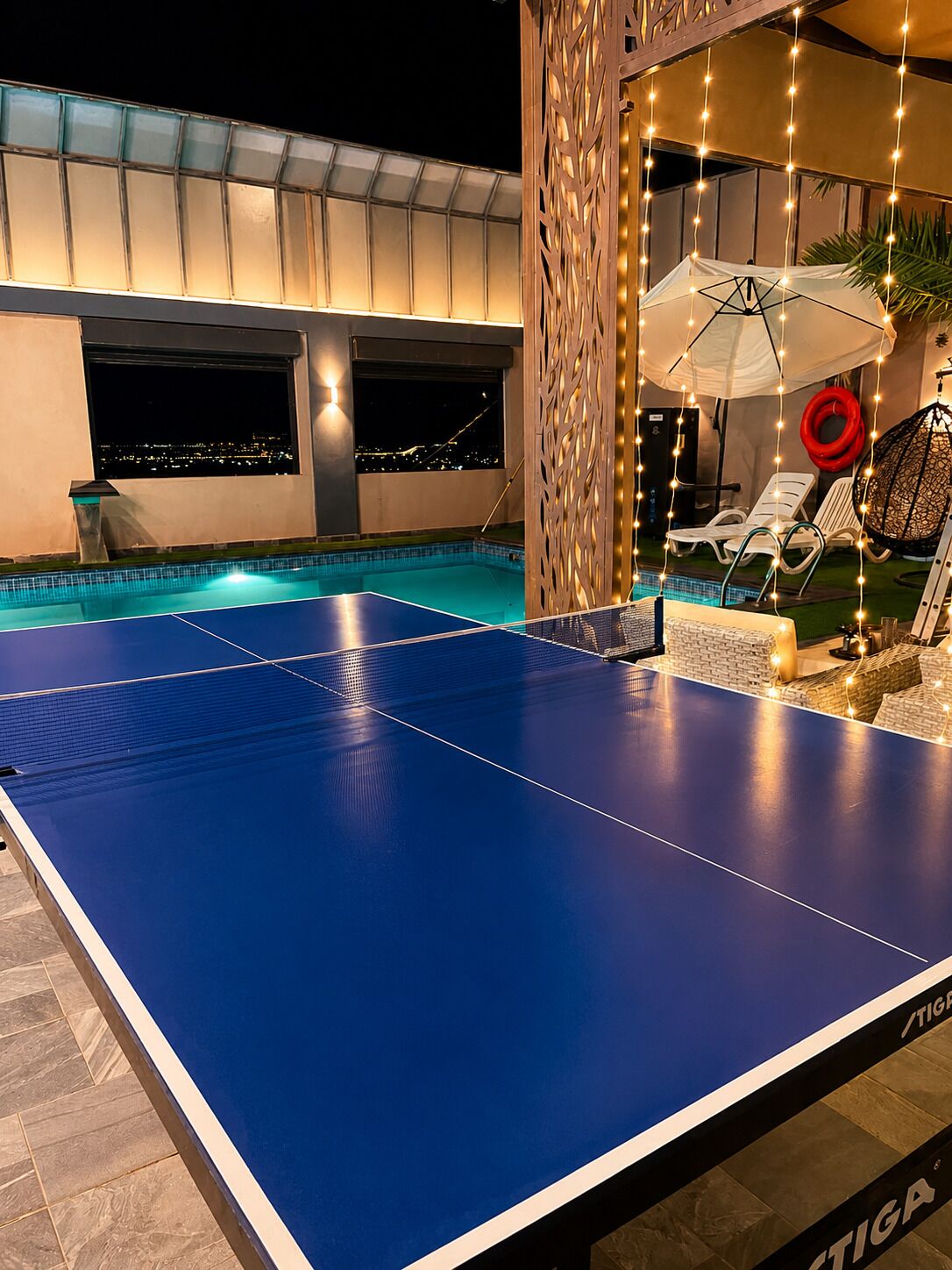 Une table de billard avec vue sur le paysage urbain illuminé la nuit, un espace de détente abrité avec des fauteuils confortables et un luminaire décoratif.