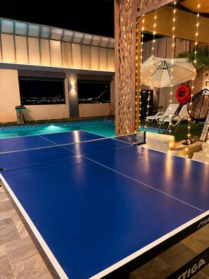 A pool table with a view of a cityscape at night, a covered seating area with lounge chairs, and a decorative light fixture. - Villa Jana chalet - Private Villa - Dead Sea - Jordan (Madaba)
