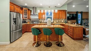 A modern kitchen with wooden cabinets, a stainless steel refrigerator, a large island with bar stools, and a tiled backsplash. - Forest Retreat • Theater • Full Spa • Speakeasy • GameRoom • MORE (East Stroudsburg)