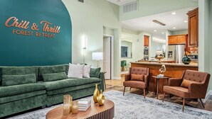 A modern living room with a green sofa, brown armchairs, and a wooden coffee table. A kitchen is visible in the background with wooden cabinets and stainless steel appliances. - Forest Retreat • Theater • Full Spa • Speakeasy • GameRoom • MORE (East Stroudsburg)