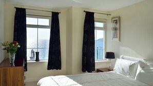 Une chambre à coucher avec un lit, une table de chevet, une lampe, une fenêtre avec des rideaux, un vase de fleurs et un tableau encadré au mur.