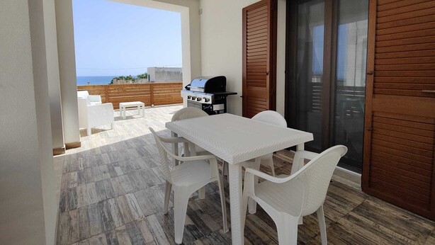Ein Balkon mit Grill, weißem Tisch und Stühlen und Blick aufs Meer.