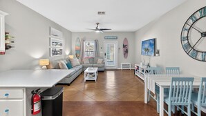 Uma sala de estar moderna com sofá, mesa de centro e área de jantar com cadeiras. Há um ventilador de teto, um relógio na parede e uma decoração com prancha de surf.