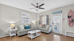 Uma sala de estar com um sofá cinza, uma mesa de centro branca e um ventilador de teto. Há uma porta com janela e uma placa com a inscrição "BEACH HOUSE".