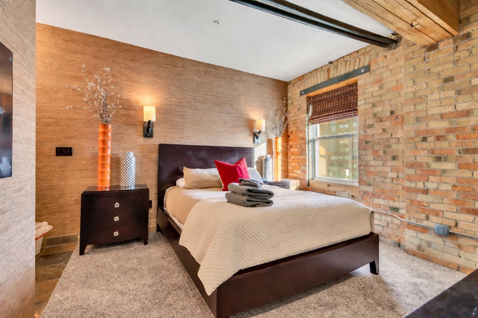 Een moderne slaapkamer met een bakstenen muur, een houten hoofdbord, een groot bed met een witte deken, een nachtkastje en een raam met jaloezieën.