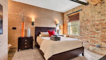 Een moderne slaapkamer met een bakstenen muur, een houten hoofdbord, een groot bed met een witte deken, een nachtkastje en een raam met jaloezieën.
