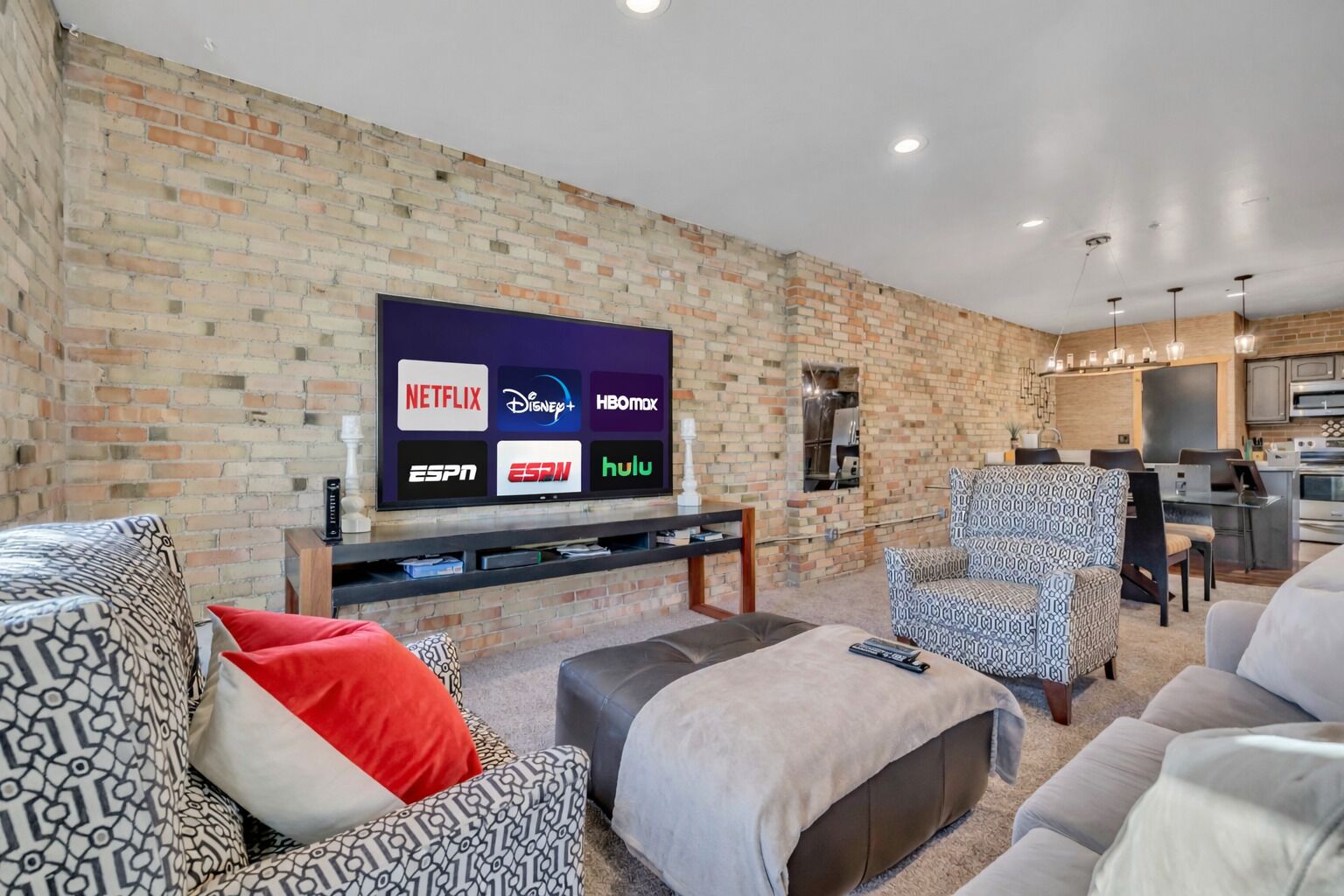 Una sala de estar con pared de ladrillo, un televisor de pantalla plana mostrando varios servicios de transmisión, y una cocina moderna al fondo.