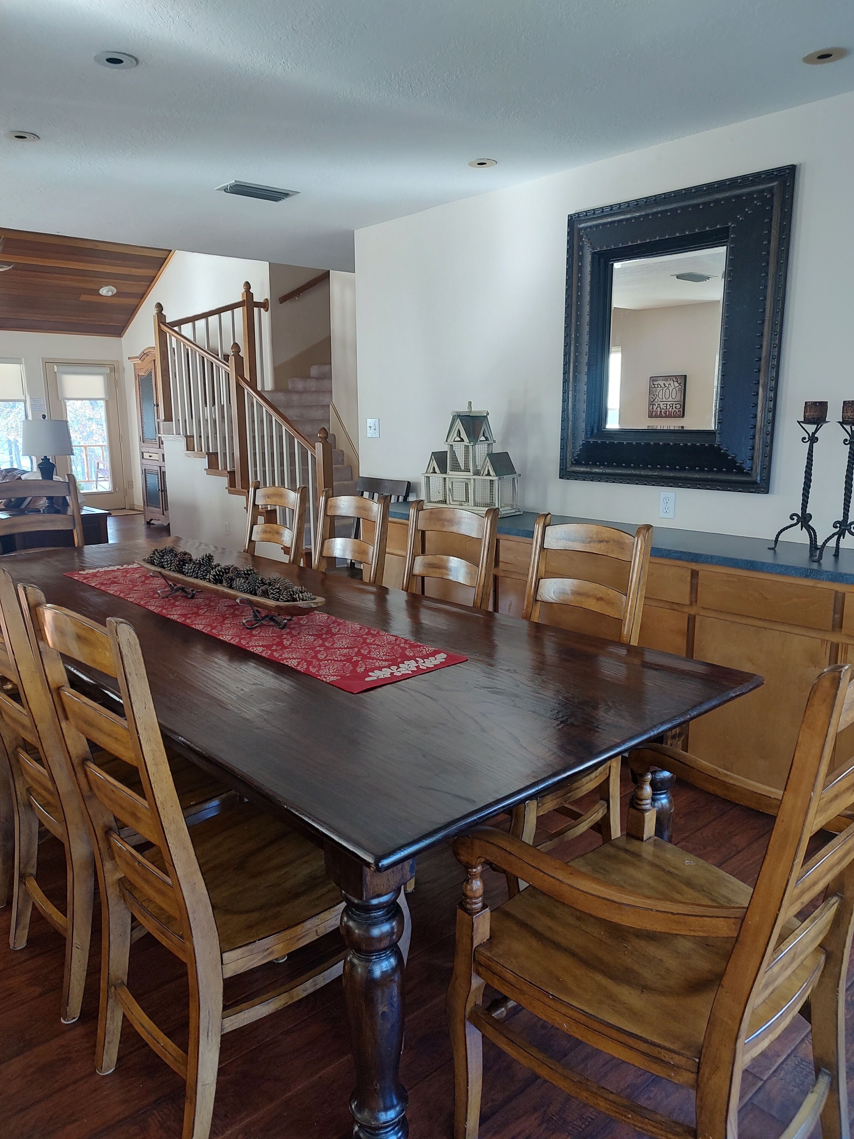 A dining area with a large wooden table, wooden chairs, a red placemat, and a decorative pine cone arrangement.