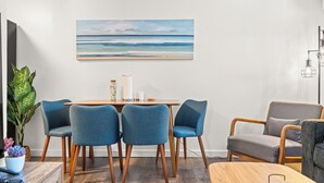 Ein modernes Wohnzimmer mit einem Holzesstisch, vier blauen Stühlen, einem grauen Sofa und einem großen Strandbild an der Wand.